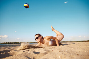 Dynamic image of young woman in motion, playing beach volleyball, hitting ball and falling down on sand. Concept of sport, active and healthy lifestyle, hobby, summertime, ad