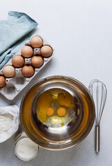 Raw eggs in stainless bowl. Carton of eggs, flour, sugar and kitchen textile napkin on concrete background. Ingredients for baking cake. Vertical photo, copy space