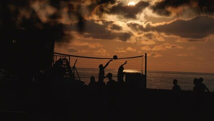 Group of people, friends or students, playing volleyball on beach by sea or ocean at sunset. Unrecognizable silhouettes of people. slow motion.