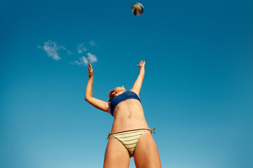 Bottom view dynamic image of young woman in swimsuit serving ball, play in g beach volleyball over blue sky background. Concept of sport, active and healthy lifestyle, hobby, summertime, ad