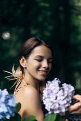 Vertical portrait of beautiful young woman standing posing in park with blooming flowers Hydrangea garden. Lady dressed summer outfit, smiling laughing having fun
