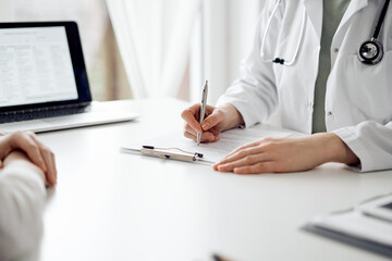 Doctor and patient sitting near each other at the white desk in clinic. Female physician is listening filling up a records form. Medicine concept
