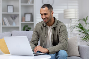 African american smiling young male student studying at home online remotely, sitting on sofa and typing on laptop