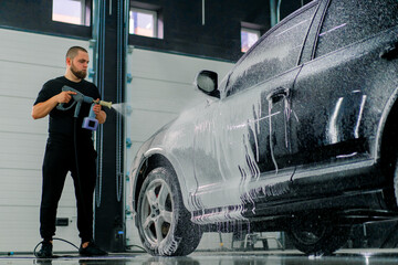 A male car wash employee applies car wash foam to a luxury black car using a spray gun in the car wash box
