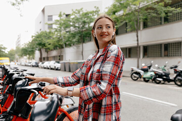 Adorable smiling girl with loose hair wearing striped shirt in wireless headphone is holding bike and preparing for cycling in the city. 