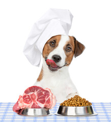 Licking lips Jack russell terrier puppy wearing a chef's hat sits with bowls with dry dog food and...