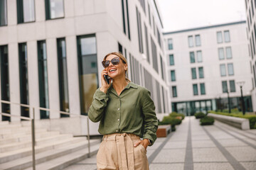 Smiling woman office worker is talking by phone while standing on modern building background 