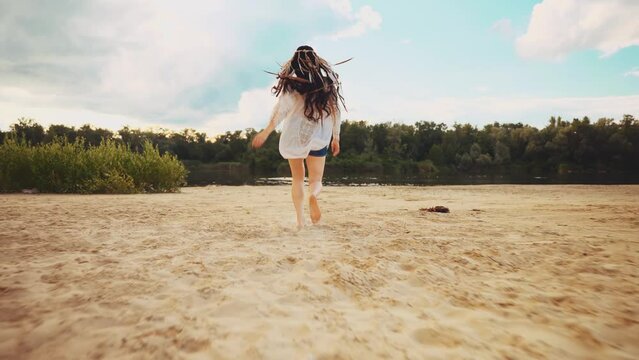 Happy woman running to river bank summer nature sand beach enjoy life freedom blue sky, back rear view long dark hair headband feather boho style. girl in white dress long shirt casual hippie clothing