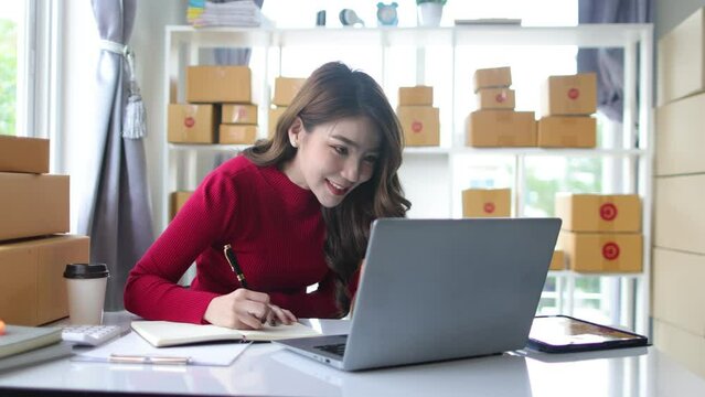 Asian woman working at home small business checking online orders to prepare boxed items for delivery to customers.