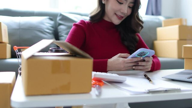 Asian woman working at home small business checking internet orders on smartphone and preparing boxed items for delivery to customers.