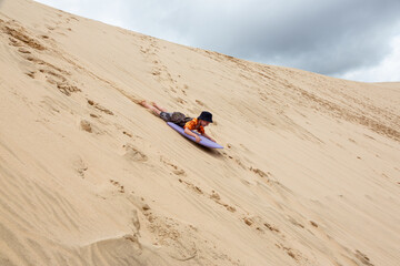 A young boy is having great fun on an adventure activity in New Zealand called sand boarding. He is...