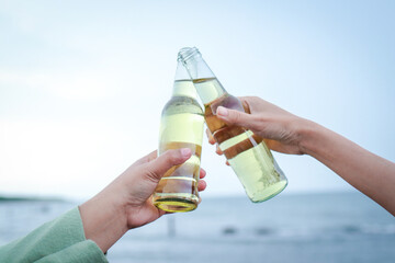 Two women partying at evening beach holding beer bottles colliding happily having fun together....