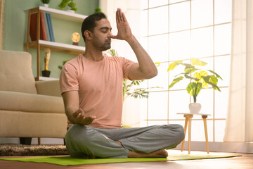 Peaceful indian man doing nostril breathing or pranayama yoga by closing eyes during morning at...