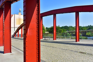 Red metal pillars of modern bridge and grey brick paved road against a blue sky. Architecture and Engineering. Poland, Poznan, June 2022.