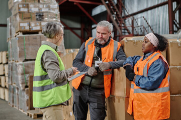 Foreman using digital tablet and holding briefing with workers during meeting in warehouse