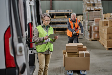 Foreman standing near the truck and using tablet pc while worker loading cargo