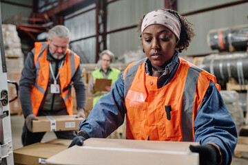 African American woman in workwear loading boxes in truck with her colleagues in background