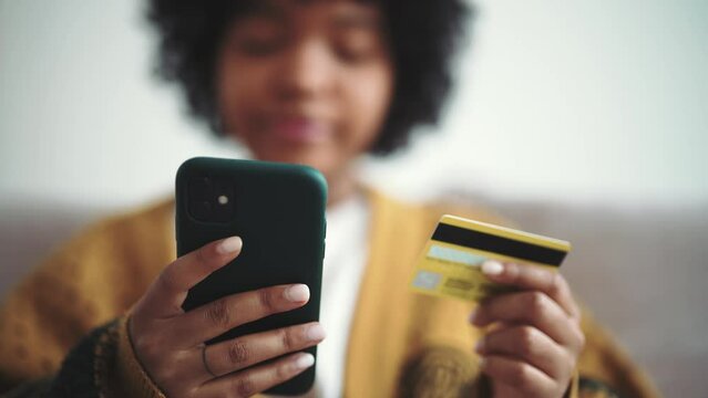 Positive african american woman doing online payment on smartphone using credit card in marketplace. Girl buys in internet at home. Online shopping, customer. Enters bank card number in online store.