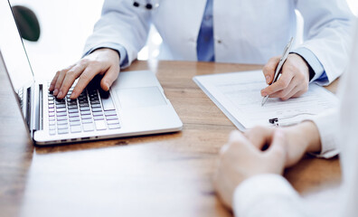Doctor and patient sitting at the wooden desk in clinic. Female physician's hands pointing into laptop computer monitor, close up. Medicine concept