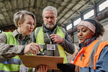 Foreman pointing at document and discussing invoice with workers during their teamwork in warehouse