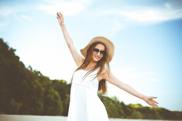 Happy smiling woman in free happiness bliss on ocean beach standing with a hat, sunglasses, and open hands. Portrait of a multicultural female model in white summer dress enjoying nature during travel