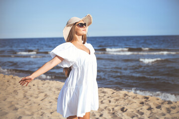 Fototapeta na wymiar Happy blonde woman is on the ocean beach in a white dress and sunglasses, open arms