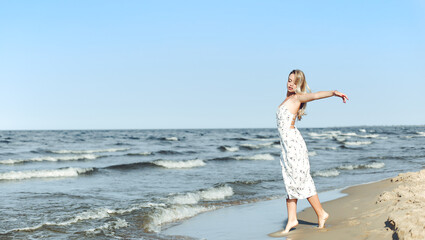 Fototapeta na wymiar Happy blonde beautiful woman on the ocean beach standing in a white summer dress, open arms