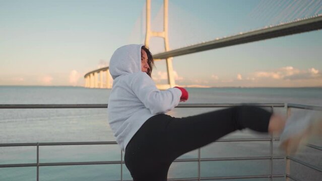 Female kickboxing fighter dressed hoodie training kicks and blocks outdoor with seascape background. Athletic boxer woman's hands protected with red boxing tapes. Camera zooms in and zoom out 