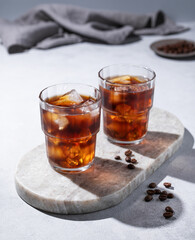 Cold brew coffee in a two glasses with ice on a marble board on a light background with coffee beans, shadows and napkin. Concept summer craft refreshing homemade drink