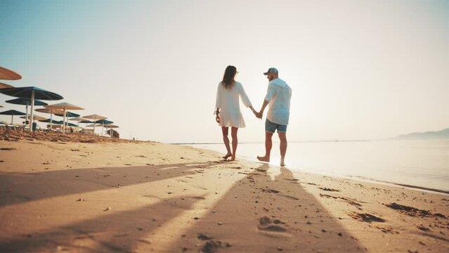 Loving couple walking along sandy ocean beach at sunny day talking enjoying time together. Man woman having romantic date, honeymoon near sea, back view. Tourism, travel family holidays concept.