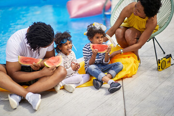 Lovely family of four sitting by the pool eating watermelon slices together. Mother and father with...