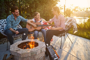 Happy girl playing guitar outdoors by the fireplace, sitting with mother and father in the backyard enjoying sunny summer day together. - 626014311
