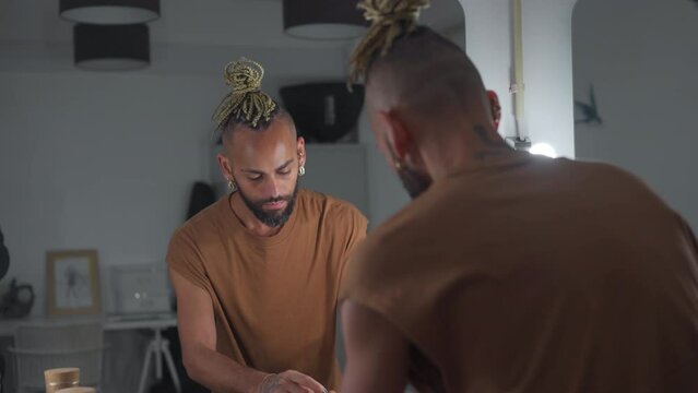 Black male gay applying make up looking mirror standing back to camera. Stylish homosexual man in dressing room doing make-up on face