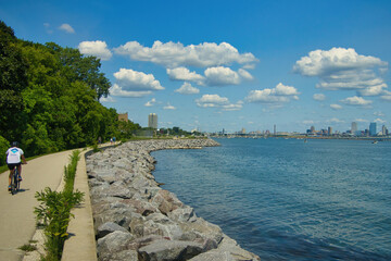 On a sunny Summer day, a cyclist, seen from behind at a distance, rides along a Lake Michigan shoreline  recreational trail approaching Downtown Milwaukee. - obrazy, fototapety, plakaty