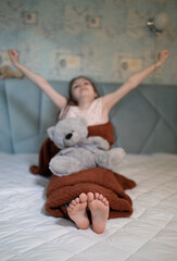 girl stretches in bed after waking up, raising her arms up and to sides. In the foreground, the bare feet of a child are in focus, a soft toy bear cub sits on its knees. Wake up early in the morning