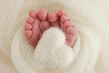 The tiny foot of a newborn baby. Soft feet of a new born in a white wool blanket. Close up of toes, heels and feet of a newborn. Knitted white heart in the legs of a baby. Macro photography