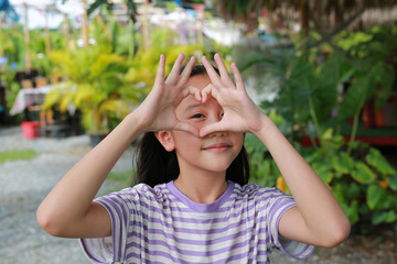 Portrait of Asian girl doing heart shape with hand and fingers smiling looking through sign in the garden.