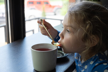 A little girl drinks tea with a teaspoon in a cafe