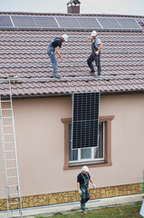 Men mounters lifting up photovoltaic solar moduls on roof of house. Electricians in helmets mounting solar panel system outdoors. Concept of alternative and renewable energy.