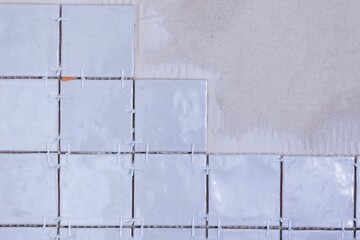 A portrait of an unfinished ceramic tile wall, with plastic white joint spacers still in the junctures and tile adhesive showing. The small tiles are rectangular and are being placed in a pattern.