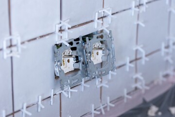 A close up portrait of two connected electric wall sockets without their protective caps in a newly tiled unfinished wall, still with the plastic white joint spacers in the junctures.