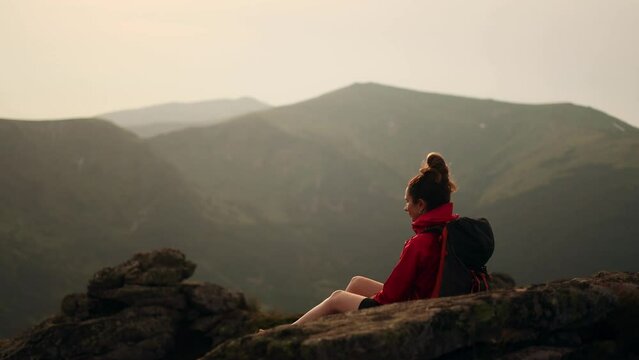 Attractive young dark haired woman hiker with backpack take a break while hiking and enjoying beautiful view of incredible mountains while sitting on the peak alone. High quality 4k footage