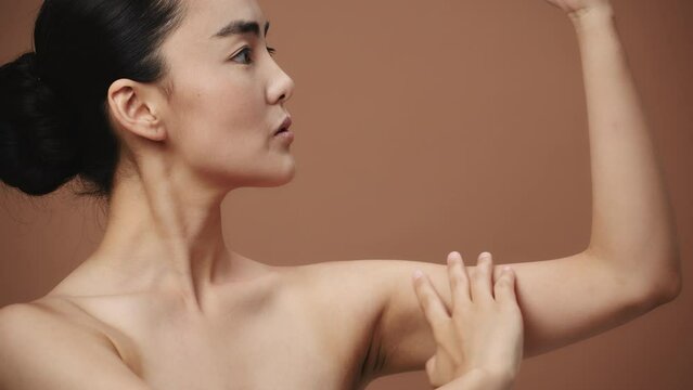 Close-up of a beauty asian young woman touches her soft skin on her bicep, on a beige background. Beautiful female model with perfect clean fresh skin. Skin care treatment concept