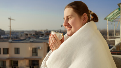 Portrait of beautiful brunette woman covered in blanket drinking coffee and looking on the city skyline