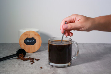 Woman's hand stirring stevia sweetener in a black coffee, grey background 