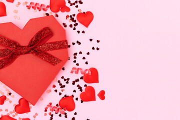 Red gift box in a heart shape tied with glittering ribbon. Festive concept with scattered confetti on a pink pastel background.