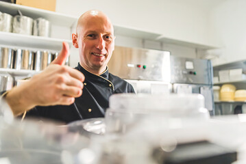 Pastry business owner with a thumbs up standing in cluttered kitchen. Bald caucasian male chef...