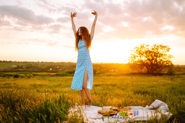 Happy woman in beautiful blue dress on picnic enjoying sunset light. Cheerful young female tourist on picnic having fun on green meadow. Concept of people, recreation, nature. Active lifestyle.