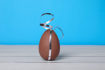 Delicious chocolate egg with silver ribbon on white wooden table against light blue background