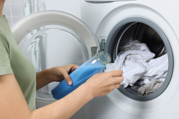 Woman pouring fabric softener into cap near washing machine with dirty clothes, closeup